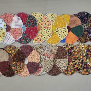 May include: A collection of circular fabric coasters arranged in two rows. Each coaster features a multi-paneled design with various autumn-themed patterns, including sunflowers, plaid, leaves, pumpkins, and solid colors in shades of orange, yellow, brown, and red.