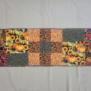 May include: A rectangular patchwork table runner featuring a variety of autumn-themed fabric squares. The design includes patterns of orange pumpkins, yellow sunflowers, dark green foliage, and multicoloured fall leaves with a frayed edge finish.