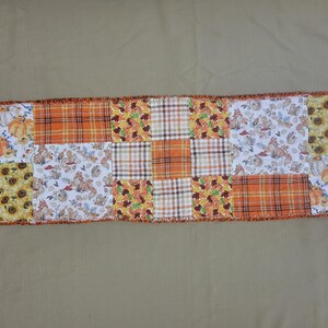 May include: A rectangular patchwork table runner featuring a variety of autumn-themed fabric squares. The design includes patterns of orange pumpkins, sunflowers, brown and orange plaid, and woodland creatures like foxes on a white background.