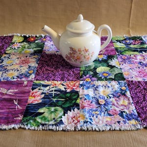 May include: A purple and green patchwork table runner with a white teapot in the centre. The runner features a floral design with water lilies and dragonflies.