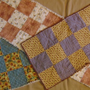 May include: Three handmade quilted table runners in different patterns. The first runner is a brown and white checkered pattern with a floral design. The second runner is a green and white checkered pattern with a sunflower design. The third runner is a purple and yellow checkered pattern with a floral design.