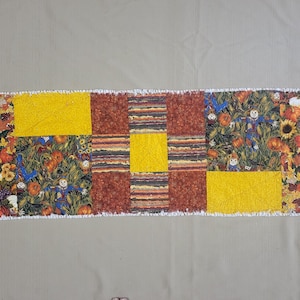 May include: A rectangular rag quilt table runner featuring a patchwork design of autumn-themed fabrics. The pattern includes panels of yellow, dark orange, and multi-coloured prints depicting sunflowers, pumpkins, and scarecrows with frayed edges.