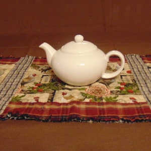 May include: A white ceramic teapot with a simple design sits on a red and white plaid table runner with a floral pattern.