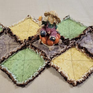 May include: A rag quilt table runner featuring diamond-shaped patches in shades of green, yellow, and brown paisley, with frayed edges. A scarecrow figurine wearing a straw hat and overalls sits atop a pumpkin in the centre of the runner.