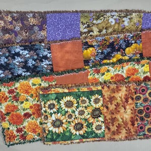 May include: A rectangular rag quilt featuring a patchwork of fabric squares with autumn-themed prints. The design includes patterns of sunflowers, orange pumpkins, fall leaves, and floral motifs in shades of purple, orange, yellow, and green.