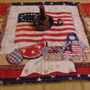 May include: A red, white, and blue patchwork quilt with a vintage American flag design. The quilt features a small doll, a heart-shaped pillow, and a candle holder.