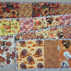 Puede incluir: Un camino de mesa tipo rag quilt con un diseño de patchwork de cuadrados de tela con motivos otoñales. Los patrones incluyen girasoles, calabazas, pavos y hojas de otoño en tonos naranja, amarillo, marrón y rojo con bordes deshilachados.