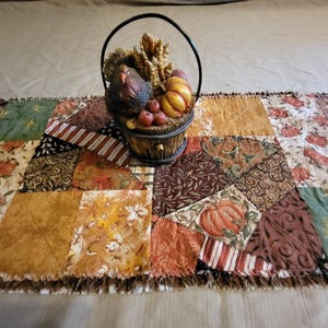 May include: A patchwork table runner with a fringed edge, featuring a variety of autumn-themed fabrics. A decorative basket with a turkey, pumpkins, wheat, and apples sits atop the runner, creating a fall harvest theme.