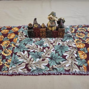 May include: A quilted table runner featuring a patchwork pattern of orange pumpkins and yellow sunflowers on a dark burgundy background, with a central panel of green and white oak leaves. A wooden block sign spells AUTUMN with a scarecrow figure.