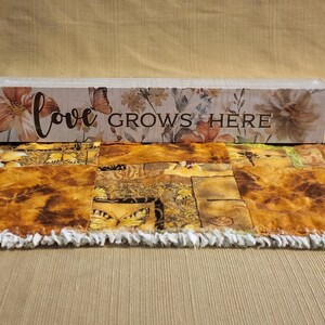 May include: A rectangular wooden sign with the words "love GROWS HERE" in black script. The sign is decorated with floral and butterfly designs in shades of peach, brown, and cream. The sign rests on a decorative table runner.