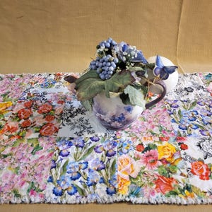 May include: A floral table runner with a patchwork design featuring various flowers in shades of pink, yellow, blue, and white. A decorative teapot with a floral design and a cluster of blue berries sits on top of the runner.