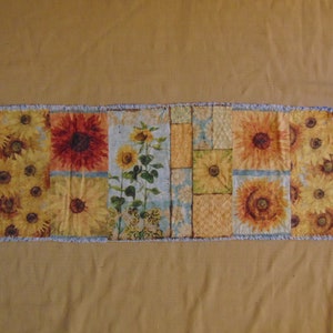 May include: A patchwork table runner with a frayed edge features a sunflower design in shades of yellow, orange, and brown. The runner is made of fabric with a textured surface.