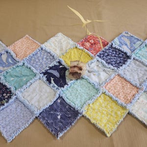 May include: A colorful, handmade quilt with a diamond pattern. The quilt features various fabrics in shades of blue, yellow, pink, and white, some with whale and floral prints. The edges are fringed, and a wooden bird sculpture is placed on top.