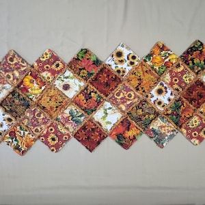 May include: A rectangular table runner constructed from numerous diamond-shaped fabric patches featuring autumn-themed prints. The patterns include sunflowers, orange pumpkins, and various fall leaves in shades of red, orange, yellow, and brown.