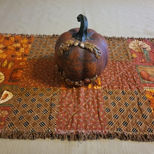 May include: A brown decorative pumpkin with a dark stem, featuring a sculpted wreath of leaves and acorns around the centre with the text Give Thanks and Be Grateful, resting on a rectangular quilted table runner with a patchwork autumn pattern.