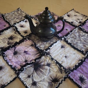 May include: A black teapot sits on a handmade quilt. The quilt is composed of square patches in white, lavender, and gray, each featuring a floral design. The edges of the quilt are trimmed with black ruffled fabric.