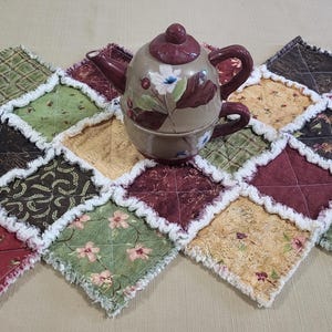 May include: A decorative teapot and teacup set in shades of brown, burgundy, and cream, with floral accents, sits atop a colorful, quilted table runner. The runner features a patchwork design with various patterned fabrics.