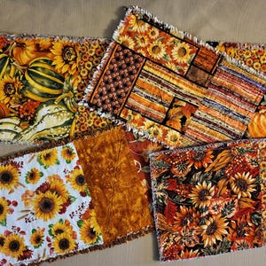 Peut inclure: Plusieurs pièces de tissu rectangulaires avec des motifs d'automne. Les motifs comprennent des tournesols, des citrouilles et des feuilles aux tons orange, jaune et marron. Les bords du tissu sont effilochés.