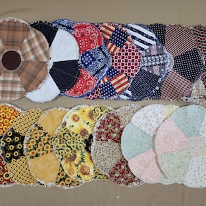 May include: A collection of handmade, circular fabric coasters. The coasters feature various patterns, including plaid, sunflowers, and patriotic designs with red, white, and blue colors. Each coaster has a frayed edge.