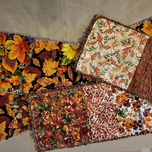 May include: A collection of autumn-themed fabric table runners and placemats. The textiles feature various fall leaf and floral patterns in shades of orange, yellow, brown, and black. The edges are finished with a fringed trim.