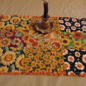 May include: A patchwork table runner with a sunflower theme. The runner is made of different fabrics with various sunflower patterns in shades of yellow, orange, and brown. The runner has a frayed edge and is decorated with a small candle holder.