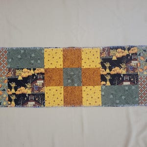 May include: A rectangular patchwork table runner featuring a variety of autumn-themed fabric squares. The design includes patterns of pumpkins, haystacks, barns, and plaid, rendered in a colour palette of orange, yellow, brown, green, and black.