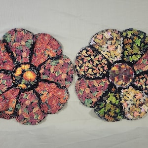 May include: Two round fabric mats shaped like flowers with frayed edges. The left mat features a pumpkin and autumn leaf print in orange, red, and brown. The right mat displays a dense pattern of colorful fall leaves on a black background.