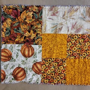 May include: A patchwork fabric item with autumn-themed patterns. The design includes squares with images of fall leaves, pumpkins, sunflowers, and floral patterns in orange, yellow, and brown. The edges have a fringed, textured finish.