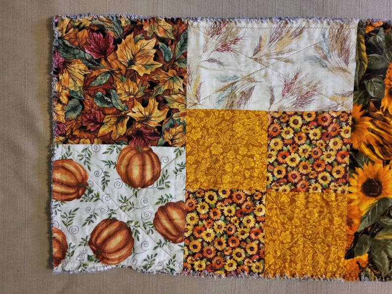 May include: A patchwork fabric item with autumn-themed patterns. The design includes squares with images of fall leaves, pumpkins, sunflowers, and floral patterns in orange, yellow, and brown. The edges have a fringed, textured finish.