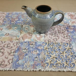 May include: A blue and tan ceramic teapot with a dark brown interior sits on a patchwork table runner. The runner features a sea life pattern with starfish, seashells, and fish in shades of blue, tan, and pink.