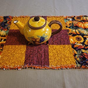 May include: A yellow ceramic teapot with a brown handle and lid sits on a patchwork table runner. The runner features squares of orange, burgundy, and black fabric with autumn-themed patterns of pumpkins, sunflowers, and leaves.