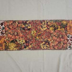 May include: A rectangular table runner featuring a patchwork design of autumn-themed fabrics. The pattern includes various prints of orange pumpkins, yellow and brown fall leaves, and dark floral motifs with frayed, textured edges on all sides.