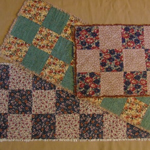 May include: Three patchwork table runners with a checkered pattern. The runners are made of different fabrics with floral prints in shades of red, blue, green, and cream. The runners have a frayed edge.