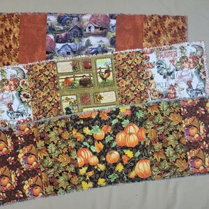 May include: Three rectangular fabric table runners featuring autumn-themed patterns. One displays a farm scene, another shows roosters and pumpkins with text, and the third features a dense print of orange pumpkins and colorful fall leaves.