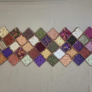 May include: A rectangular table runner constructed from numerous diamond-shaped fabric patches. The patches feature a variety of colours including purple, orange, green, yellow, and brown, with diverse floral, striped, and geometric patterns.