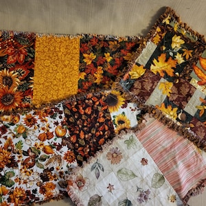 Peut inclure: Un assortiment de carrés de tissu aux bords effilochés, sur le thème de l'automne. Les carrés présentent des motifs de tournesols, de feuilles et de motifs floraux dans des tons orange, jaune, marron et crème. L'ensemble est rustique et saisonnier.