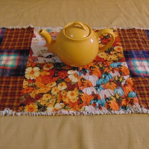 May include: A yellow ceramic teapot sits on a patchwork table runner. The runner features a variety of fall-themed fabrics, including plaid, floral, and pumpkin patterns. The edges of the runner have a fringed trim, adding a rustic touch.