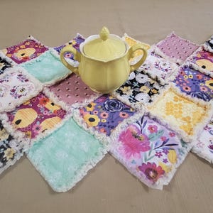 May include: A yellow ceramic sugar bowl with a lid sits on a patchwork quilt. The quilt is made of various floral patterned squares in colors like purple, pink, yellow, and white. The quilt has a fringed edge.