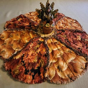 May include: A decorative table topper with a fall theme, featuring a central figurine. The topper is made of fabric in shades of orange, brown, and yellow, with floral and leaf patterns. The figurine has a squirrel holding a pumpkin with the word "THANK" on it.