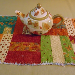 Puede incluir: Una tetera de cerámica con diseño de hojas sobre un camino de mesa de patchwork. La tetera tiene una base beige con detalles de hojas marrones y un asa tejida. El camino de mesa presenta telas con temática otoñal en naranja, rojo, verde y beige.
