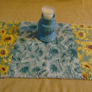 May include: A blue and yellow table runner with a sunflower pattern. A blue ceramic candle holder with a white candle is in the center of the runner.
