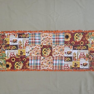 May include: A rectangular fabric table runner featuring a patchwork design of autumn-themed patterns including sunflowers, pumpkins, mushrooms, plaid squares, and red trucks with the text Thankful. The edges are finished with a frayed orange fringe.
