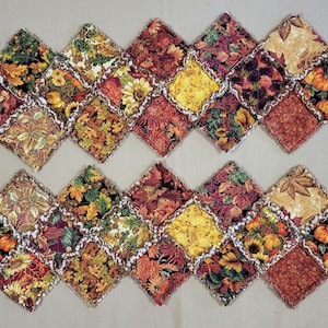 May include: Two rectangular fabric table runners featuring a patchwork design of diamond-shaped squares. The fabric displays autumn-themed patterns including orange pumpkins, yellow sunflowers, and various fall leaves in shades of red, gold, and brown.