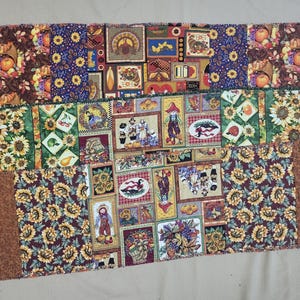May include: A rectangular fabric table runner featuring a patchwork design with three distinct horizontal sections. The patterns include sunflowers, pumpkins, cornucopias, scarecrows, and autumn harvest motifs in shades of orange, brown, green, and yellow.