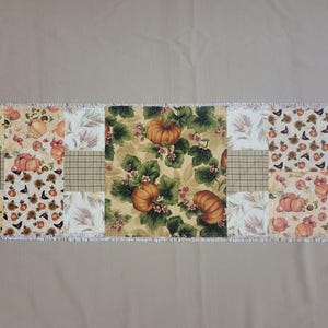 Peut inclure: Un chemin de table patchwork rectangulaire composé de divers carrés de tissu sur le thème de l'automne. Le motif présente des citrouilles orange, des feuilles vertes, des tournesols, des épis de blé et un carreau marron, avec un bord effiloché blanc.