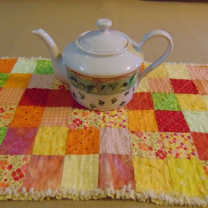 Puede incluir: Una tetera blanca con un diseño floral se encuentra sobre un camino de mesa de patchwork colorido. El camino de mesa está hecho de cuadrados de tela en tonos de amarillo, naranja, rosa y verde.