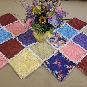 Peut inclure: Une nappe matelassée colorée avec un motif patchwork. Les carrés sont dans les tons de rouge, bleu, rose, jaune et violet. Le chemin de table est décoré d'un vase de fleurs.