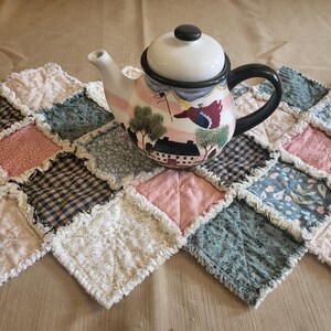 May include: A decorative teapot with a black handle and lid, featuring a painted scene of a house and trees. It sits on a patchwork quilt with pink, blue, and black squares. The quilt has a ruffled edge.
