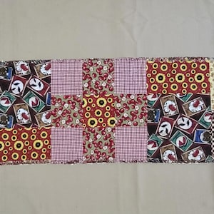May include: A rectangular patchwork table runner featuring a variety of fabric squares. The patterns include red and white gingham, sunflowers on a red background, cherries on a green background, and a print of various fruit-themed square labels.