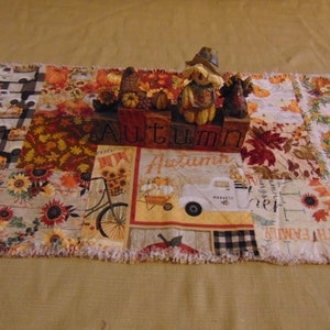 May include: A patchwork table runner with a fall theme. The runner features various patterns including sunflowers, pumpkins, and harvest trucks. A wooden "AUTUMN" block and a scarecrow figurine are placed on top.